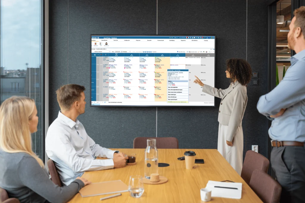 Een groep mensen in een vergaderruimte kijkt naar een groot scherm met informatie over een urenregistratiesysteem.